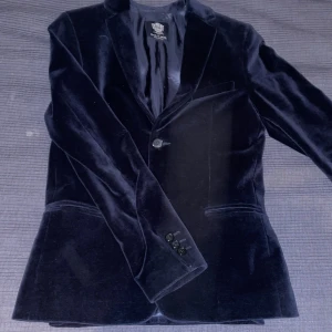 Mörkblå sammetskavaj från Dal Lago - Elegant mörk blå kavaj i sammet från Dal Lago, tillverkad i Italien. Kavajen har en klassisk design med två knappar framtill och en slät rygg. Perfekt för formella tillfällen. Storlek 13 år 158. Ny pris 5500