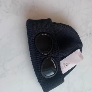 Svart mössa från C.P. Company - Snygg svart mössa från C.P. Company med unika glasögondetaljer framtill. Mössan är ribbstickad och har en klassisk design som passar perfekt för kyligare väder. Perfekt för vintern. 