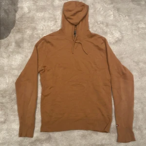 Brun hoodie från Tommy Hilfiger - Säljer en snygg brun hoodie från Tommy Hilfiger i storlek M. Den har en klassisk design med huva och dragsko, samt ribbade muddar vid ärmslut och nederkant. Perfekt för en avslappnad stil.
