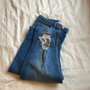 Blå jeans med slitningar - Säljer ett par snygga blå jeans med slitningar på knäna. Perfekta för en avslappnad stil. De har en klassisk femficksdesign och knappgylf.
