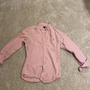 Rosa skjorta från Ralph Lauren - Säljer en stilren rosa skjorta från Ralph Lauren i slim fit. Skjortan har långa ärmar och knappar framtill samt en broderad logotyp på bröstet. Perfekt för en elegant look.