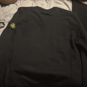Svart tröja från Stone Island - Säljer en svart tröja från Stone Island med deras ikoniska märke på ärmen. Tröjan har en klassisk design med rund hals och långa ärmar. Perfekt för en stilren och avslappnad look.