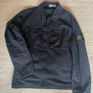 Svart overshirt från Stone Island - Snygg svart overshirt från Stone Island med dragkedja och klassisk krage. Jackan har långa ärmar och två bröstfickor med lock. Perfekt för en stilren look.
