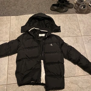 Svart pufferjacka från Calvin Klein - Säljer en snygg svart pufferjacka från Calvin Klein strl M med huva och dragkedja. Jackan är använd ca 10 gånger.  Perfekt för kyligare dagar. Pris kan diskuteras vid snabb affär!