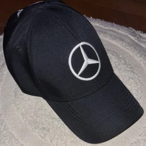 Sommar25. Svart keps med Mercedes AMG Petronas broderi - Snygg svart keps med broderad Mercedes-logga och AMG Petronas Formula One Team text. Kepsen har även gröna detaljer från Monster Energy. Den är inköpt i Nice på F1 butiken för 79 €, haft den ett par tillfällen när jag hade mercedes. Perfekt för racingfans! Snygga detaljer som inte finns på billigare kepsar, Denna hade Hamilton en exakt likadan på sig-23.