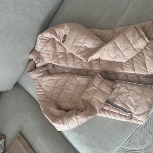 Beige quiltad jacka från Barbour - Säljer en snygg beige quiltad jacka från Barbour. Jackan har en dragkedja framtill och är perfekt för kyligare dagar. Den har en stilren design med långa ärmar och en bekväm passform.
