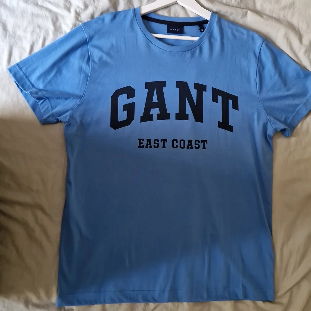 Säljer en snygg blå t-shirt från GANT med texten 'EAST COAST' tryckt på framsidan. Den är i storlek L och gjord av mjuk bomull, perfekt för en avslappnad stil. Passar bra till både jeans och shorts! Helt NY, aldrig använd!. T-shirts.