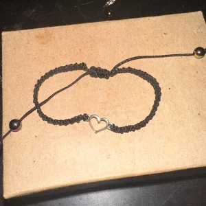 Svart armband med hjärtdetalj - Säljer ett snyggt svart armband med en söt hjärtdetalj i silver. Armbandet är flätat och justerbart med små pärlor i ändarna. Perfekt för att ge en enkel men stilren look till din outfit!