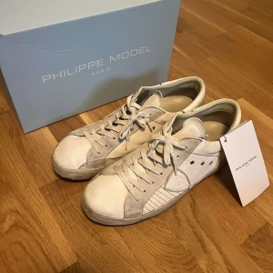 Vita sneakers från Philippe Model - Säljer ett par snygga vita sneakers från Philippe Model. Skorna har en stilren design med snörning och detaljer i mocka. De är i bra skick och perfekta för både vardag och fest. Kommer med originalkartong.