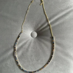 Färgglatt halsband med stenar - Säljer ett supervackert halsband med en kedja i guldton. Halsbandet är dekorerat med små, glittriga stenar i olika färger som blå, lila och svart. Perfekt för att ge lite extra färg och stil till din outfit. Det har en enkel och säker låsning. 