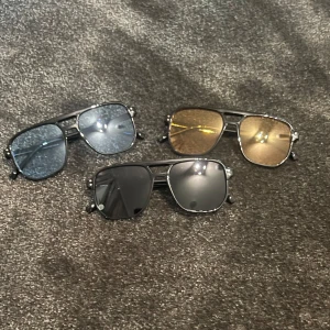 Tre par solglasögon i olika färger - Säljer tre par snygga solglasögon med metallbågar. Ett par har blå linser, ett annat par har guldiga linser och det sista paret har svarta linser. Alla har en klassisk aviator-stil med en modern twist. Perfekta för att ge din outfit en cool touch!