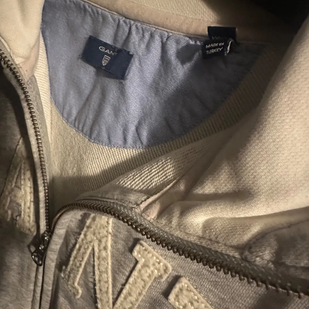 Säljer en snygg och bekväm grå hoodie från GANT. Den har en dragkedja framtill och stora bokstäver i vitt på bröstet. Perfekt för en avslappnad stil och passar till alla tillfällen. Den är i bra skick och redo för en ny ägare! (äkta). Neuletakit & Villapaidat.
