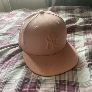 Rosa keps från New Era - Säljer en snygg rosa keps från New Era med en broderad logga framtill. Kepsen har en klassisk design med en rak skärm och är perfekt för både vardag och sportiga aktiviteter. Den är i bra skick och redo för nya äventyr!
