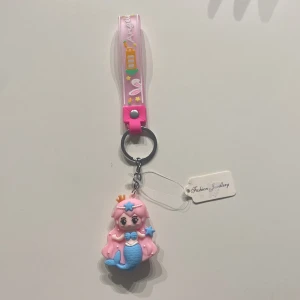 Nyckelring med sjöjungfru från Fashion Jewellery - Supergullig nyckelring med en sjöjungfrufigur i rosa och blått. Sjöjungfrun har en liten krona på huvudet och detaljer som små pärlor i håret. Nyckelringen har ett rosa band med texten 'Angel' och små dekorationer som stjärnor och en morot. Perfekt accessoar för att ge lite magi till dina nycklar eller väska!