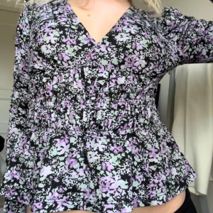 Blommig blus med volanger - Säljer en superfin blommig blus med volanger. Den har en v-ringad hals och långa ärmar. I midjan drar sömmarna åt och ger en jätte snygg figursydd passform ❤️Blusen är i en härlig mix av svart, lila och vit med ett blommigt mönster. Perfekt för både vardag och fest!