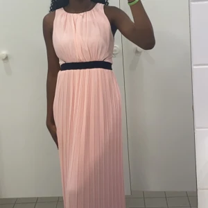Ljusrosa långklänning från Vila - Säljer en superfin ljusrosa långklänning från Vila. Den har en elegant plisserad design och en svart midjeband som ger en snygg kontrast. Klänningen är ärmlös och har en liten öppning i ryggen med dragkedja. Perfekt för sommarens fester eller en speciell kväll! 🌸