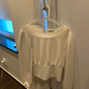 Vit ribbad topp - Säljer en snygg vit ribbad topp med långa ärmar. Toppen har en elegant design med en liten öppning i ryggen och en croppad passform. Perfekt för både vardag och fest!