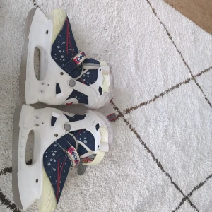 Vita och blåa barnpjäxor med stjärndetaljer - Säljer ett par coola barn skridskor i vitt och blått med röda stjärndetaljer. De har ett snöflingemönster och är utrustade med justerbara spännen för perfekt passform. Insidan är fodrad med mjukt material för extra värme. Perfekta för vinterns äventyr i snön!