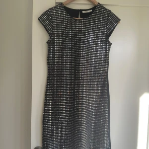 Glittrig klänning från KappAhl - Säljer en superfin glittrig klänning från KappAhl i storlek 170. Klänningen är svart med ett mönster av glittriga paljetter som ger en festlig look. Den har korta ärmar och en dragkedja i ryggen för enkel påklädning. Perfekt för nyårsfesten eller andra speciella tillfällen!