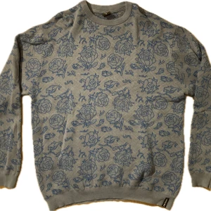 Grå tröja med blått blommönster från Santa Cruz - Säljer en snygg grå tröja från Santa Cruz med ett coolt blått blommönster över hela. Den är perfekt för både höst och vinter med sin sköna och varma känsla. Tröjan har långa ärmar och en ribbad krage som ger en stilren look. Passar perfekt för en avslappnad stil eller när du vill sticka ut lite extra! Tyvärr har den galgmärken, utöver det har den burits väldigt sällan och är i toppskick.