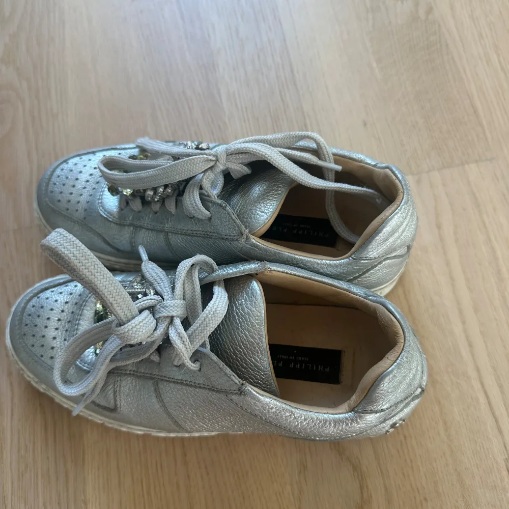 Säljer ett par coola silvriga sneakers från Philipp Plein. De har en glittrig look med pärldetaljer på snörningen och en präglad logga på sulan. Skorna är i skinn och har en normal passform. Perfekta för att ge din outfit en extra touch!. Kengät.