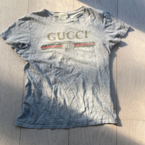 Grå t-shirt från Gucci - Säljer en snygg grå t-shirt från Gucci med det klassiska logotyptrycket i guld och de ikoniska gröna och röda ränderna. T-shirten är kortärmad och i bomull i storleken XXS. Kan sänka pris vid snabbt köp☺️