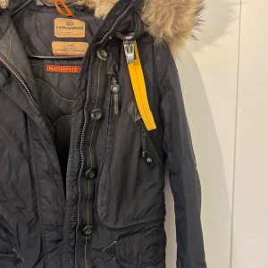 Svart vinterjacka från Parajumpers - Säljer en snygg och varm svart vinterjacka från Parajumpers. Jackan har en fluffig pälsfodrad huva och är utrustad med både dragkedja och knappar för extra skydd mot kylan. Den har flera praktiska fickor och en cool gul detalj vid halsen. Perfekt för kalla vinterdagar!