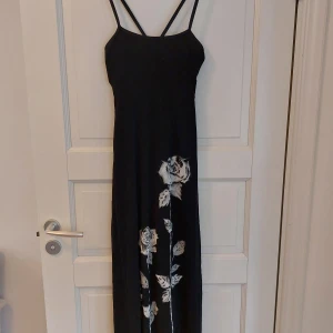 Festlig långklänning - Svart långklänning med tunna band som korsas i ryggen. Motiv av tre vita rosor i olika höjd, dessa har dekorationer av små paljetter/pärlor. Lapp bortklippt, okänt märke. Uppskattad storlek M / 40.