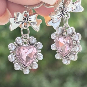 Söta örhängen med hjärtformade stenar och pärlor - Dessa örhängen är verkligen bedårande! De har en hjärtformad rosa sten i mitten, omgiven av små vita pärlor. Ovanför hjärtat finns en söt liten rosett i silverfärg. Perfekta för att ge en romantisk touch till vilken outfit som helst! Ny 