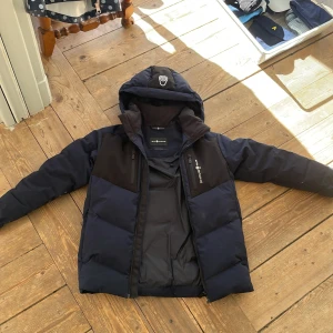 Mörkblå dunjacka från Sail Racing - Snygg mörkblå dunjacka från Sail Racing i superfint skick. Fem fickor med dragkedja. Funkar perfekt som vinterjacka och som skidjacka. Har bara använts av ett barn i en säsong. Små märken på undersidan av båda ärmarna (se bild) i övrigt som nyskick. 