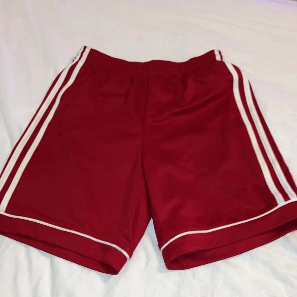 Säljer ett par röda Adidas shorts i mycket bra skick. De har de klassiska tre vita ränderna längs sidorna och är gjorda i ett bekvämt polyester material. Perfekta för träning eller en avslappnad dag. Passar bra till sommarens alla aktiviteter! 🔥. Shorts.