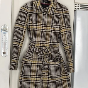 Rutig kappa från Riley De Luxe i ull  - Säljer en superfin rutig kappa från Riley De Luxe i storlek S. Den har ett klassiskt hundtandsmönster i svart, vitt och gult. Kappan är långärmad och har ett bälte i midjan för en snygg passform. Perfekt för höst och vår!