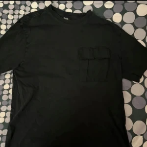 Svart t-shirt från Black Squad - Säljer en svart t-shirt från Black Squad i storlek S. Den har en cool oversized passform och en praktisk ficka på bröstet. Perfekt för en avslappnad stil. Tillverkad i Bangladesh.