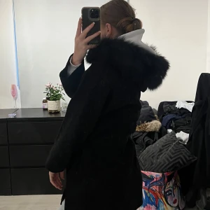 Svart pälskappa - Säljer en elegant svart kappa med stor pälskrage. Perfekt för vintern med sin varma och stiliga design. Den har långa ärmar och ett bälte i midjan för en snygg passform. Passar både till vardags och festliga tillfällen. 🖤