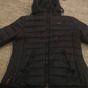 Svart pufferjacka från Navahoo - Säljer en svart pufferjacka från Navahoo i mycket bra skick. Jackan har en dragkedja framtill och två praktiska fickor med dragkedjor. Den är perfekt för kyliga dagar och har en mysig huva. Passar både till vardags och lite mer avslappnade tillfällen.