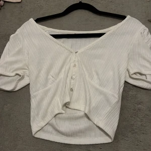 Vit ribbad topp med knappar - Säljer en söt vit ribbad topp med korta ärmar och knappar framtill. Toppen har en offshoulder-design som ger en avslappnad och stilren look. Perfekt för sommardagar eller en avslappnad festkväll. Passar bra till både jeans och kjol!