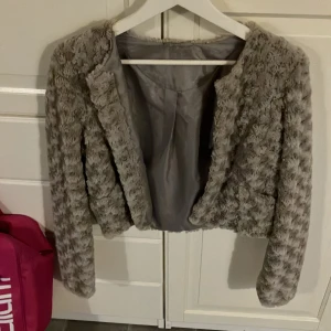 Grå pälsjacka - Säljer en supermjuk och fluffig grå pälsjacka i croppad stil. Perfekt för att hålla sig varm och snygg under kyliga höst- och vinterdagar. Jackan har långa ärmar och en stilren design utan knappar eller dragkedja. Passar perfekt till både vardag och fest!