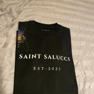 Svart t-shirt från Saint Salucci - Snygg svart t-shirt från Saint Salucci med tryck 'EST. 2021' på framsidan. Den har en cool färgexplosion på axeln och en gul etikett med knappdetalj längst ner. Perfekt för en casual look!