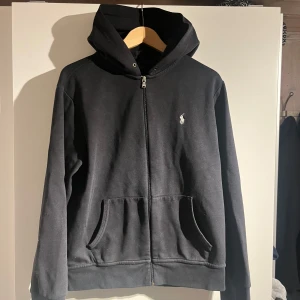 Svart hoodie från Ralph Lauren - Säljer en stilren svart hoodie från Ralph Lauren i storlek M. Den har en dragkedja framtill och en liten broderad logga på bröstet. Perfekt för både vardag och chill