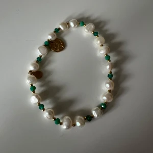 Pärlarmband med gröna detaljer från By Jolima - Snyggt armband med vita pärlor och små gröna pärlor emellan. Armbandet har också guldfärgade detaljer och en liten medaljong. Perfekt för att piffa upp vilken outfit som helst! Nyskick!!