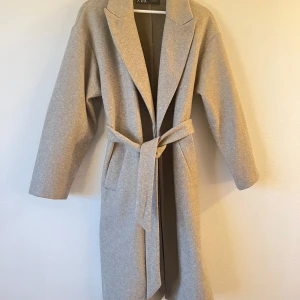 Beige kappa från Zara - Säljer en superfin beige kappa från Zara i mycket bra skick. Den har en klassisk design med knytbälte i midjan och långa ärmar. Perfekt för höst och vår när du vill vara både stilren och varm. Passar till både vardag och fest!