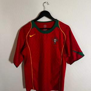 Säljer en röd Portugal fotbollströja från 2004 Nike i bra skick. Tröjan har korta ärmar och gröna detaljer vid kragen och ärmsluten. Det finns gula linjer längs sidorna och Portugals emblem på bröstet. Perfekt för fotbollsfans! ⚽