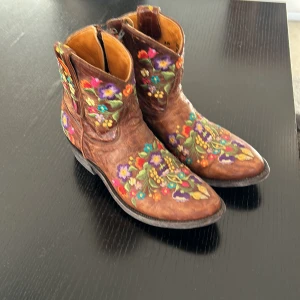 Bruna broderade boots - Säljer ett par unika Mexicana  bruna boots i skinn med färgglatt blommigt broderi. De har en dragkedja på sidan för enkel på- och avtagning. Perfekta för att ge en bohemisk touch till din outfit. Tillverkade i Mexiko och i mycket bra skick.