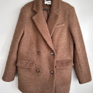Brun kappa från NA-KD - Säljer en superfin brun kappa från NA-KD i mycket bra skick. Den är långärmad och har en dubbelknäppt design med stora knappar. Perfekt för höst och vinter, och den är gjord i ett mysigt ullmaterial . Storleken är 36. Passar både till vardags och lite finare tillfällen.