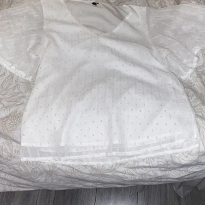 Vit blus med v-ringning - Säljer en söt vit blus med v-ringning och korta ärmar. Blusen har ett diskret prickigt mönster och är perfekt för både vardag och fest. Den är i mycket bra skick och passar till jeans eller kjol för en avslappnad men stilren look.