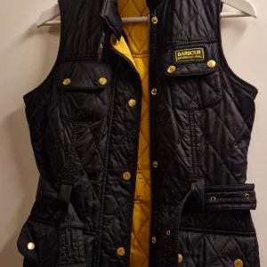 Svart quiltad väst från Barbour - Säljer en snygg svart quiltad väst från Barbour i storlek EUR 34. Västen har gul insida och detaljer som knappar och bälte i midjan. Perfekt för höst och vår, med en stilren look som passar både till vardags och fest.