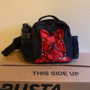 Svart och röd midjeväska från Hike - Säljer en praktisk svart och röd midjeväska från Hiker. Perfekt för vandring eller utflykter. Väskan har en justerbar rem och flera fack, inklusive en hållare för vattenflaska. Den är i mycket bra skick och redo för nya äventyr! Vattenflaskan ingår.