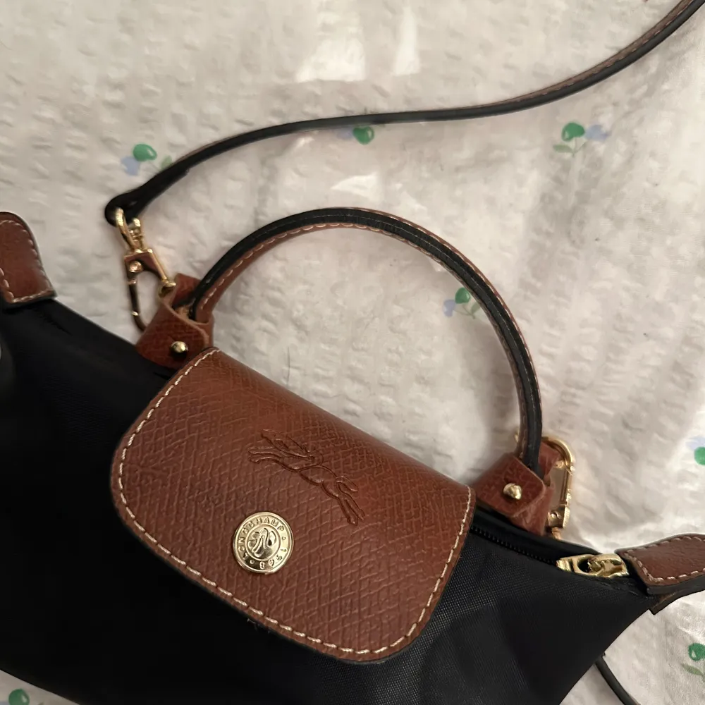 Snygg och praktisk svart axelväska från longchamp med bruna detaljer. Väskan har ett elegant märke på framsidan och en dragkedja för säker förvaring. Perfekt för både vardag och festliga tillfällen. Den justerbara axelremmen gör den bekväm att bära.. Väskor.