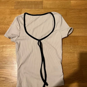 Vit ribbad topp med svart knytband - Säljer en superfin vit ribbad topp med svart kant och knytband framtill. Den är helt ny och är oanvänd eftersom den är för liten för mig