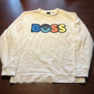 Vit sweatshirt från BOSS {M} - Säljer en vit sweatshirt från BOSS i mycket bra skick (har typ aldrig används). Färgglatt tryck med bokstäverna BOSS i blått, svart, orange och gult på framsidan. Tröjan är från johnells och går för nypris på 1599. 